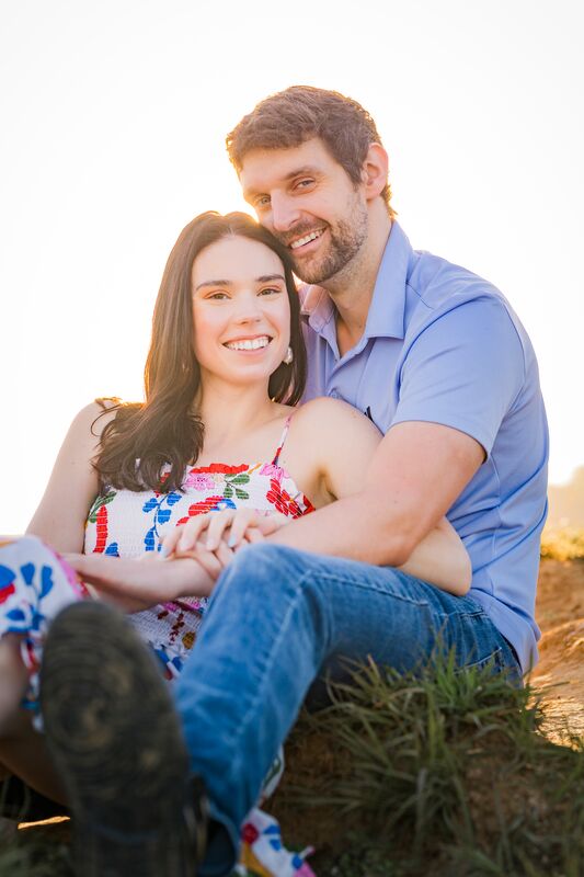 Ryan and Theresa sitting close on a grassy, sunlit hillside, both smiling at the camera