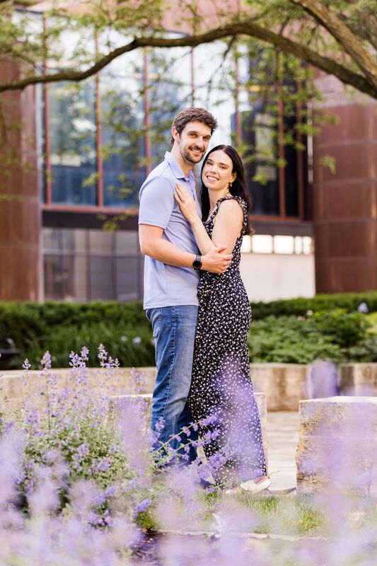 Theresa and Ryan standing close together in a courtyard, purple flowers in the foreground